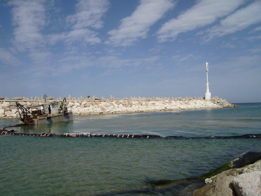 Uitbaggeren van de haveningang. Die is bijna helemaal dichtgeslibd op een smalle doorvaart na, gemarkeerd met een rode boei (links onder het havenhoofd, als je goed kijkt)